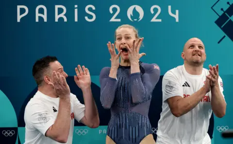 Getty Images Bryony Page, in a blue sparkly leotard, reacts in shock with her hands raised to her face and her mouth open in surpise. She is stood between her coaches, two men in white t-shirts, who are clapping.