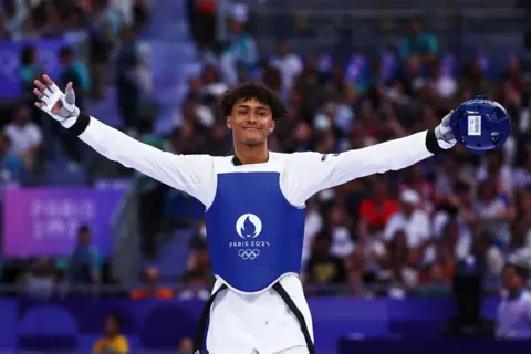 Getty Images Caden Cunningham stands with his arms stretched wide, a blue helmet in one hand. He is smiling and wearing all white with a blue vest with the Paris 2024 logo on. The crowd behind him is blurred.