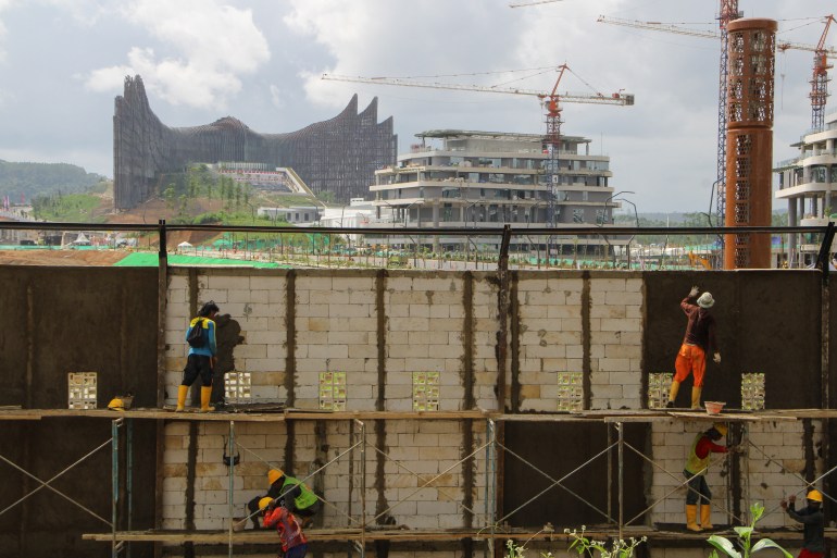 People working to build a wall. The presidential palace and other partly completed buildings are behind.