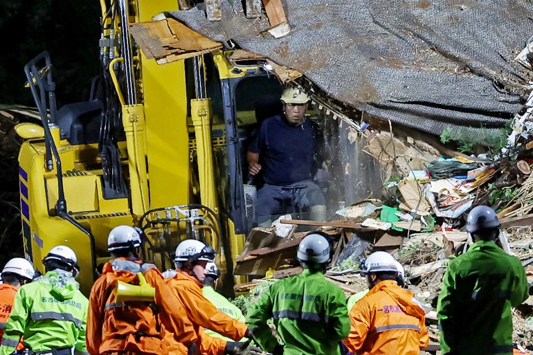 Rescuers at the site of a house that was buried in a landslide. One man is in an excavator moving debris. Others are searching the ruins