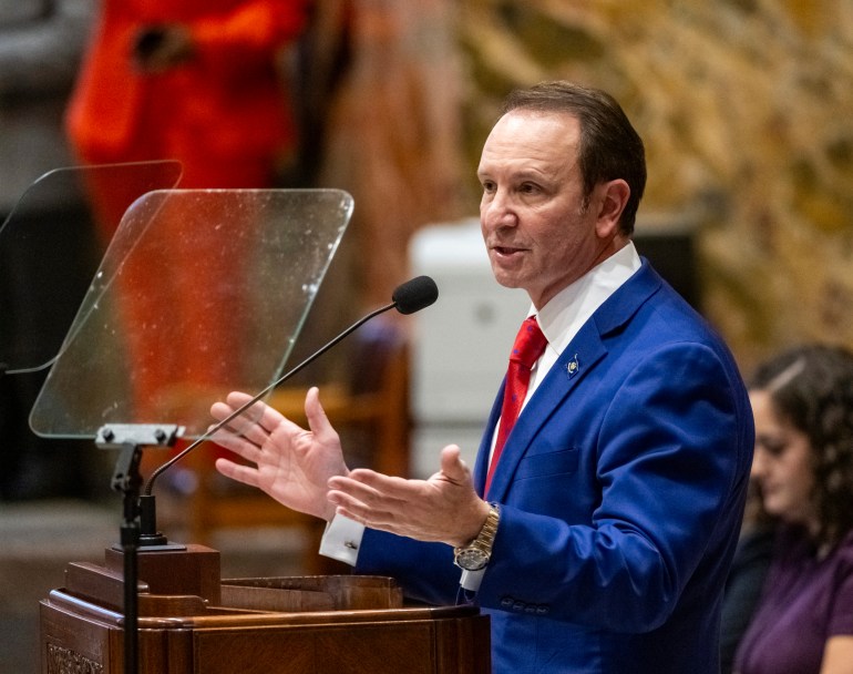 Jeff Landry speaks behind a wooden podium that has a clear shield in front of its microphone. He wears a blue suit and red tie.