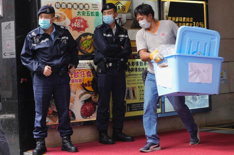 Uniformed police outside Stand News office during a raid. A man is carrying a large blue container out of the building