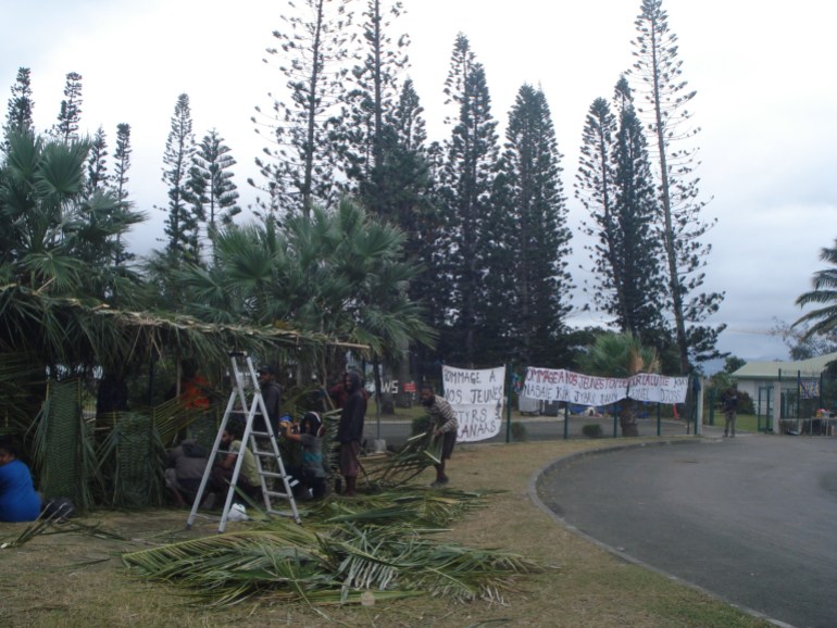 An encampment by the road side in Noumea. There are palm fronts on the roof and some people are weaving walls from the fronds There are signs in French behind them in honour of the young kanaks killed in May's violence.