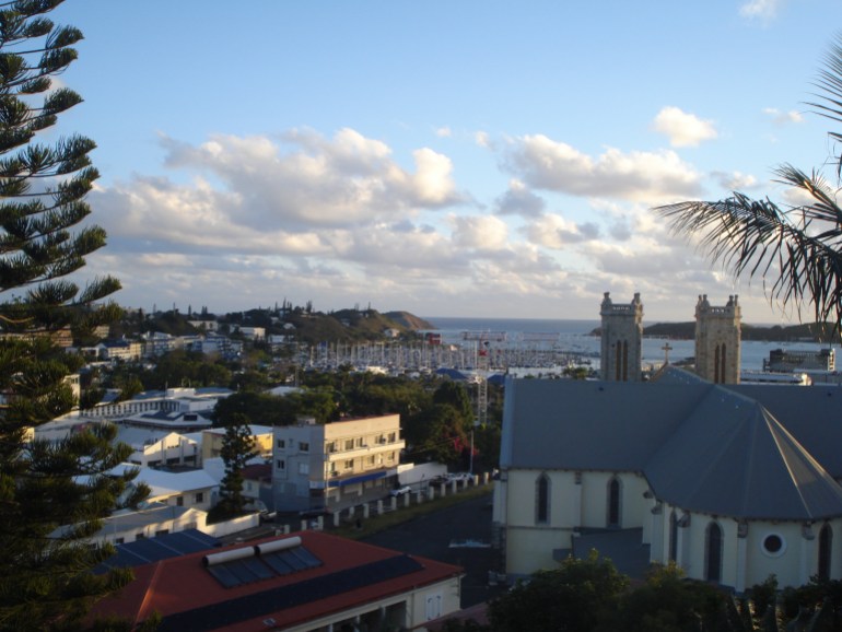 A view of Noumea from a hill above the city. The sky is blue with scattered clouds. It looks tranquil 