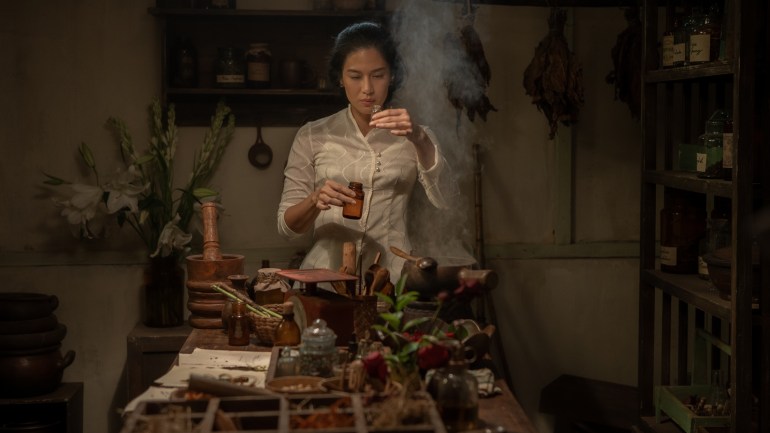 Dian Sastrowardoyo as Dasiyah in Cigarette Girl. She is wearing a cream coloured top and brown batik sarong. She is inside a laboratory and holding two bottles. Steam is rising from a bowl on the table in front of her. There are flowers behind. She is concentrating.