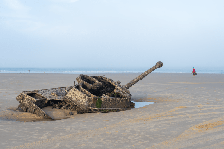 An old tank submerged in sand on a Kinmen beach. There is a person walking in the distance.