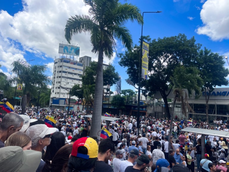 Crowds gather in an open square in Caracas where palm trees and high-rises are visible.