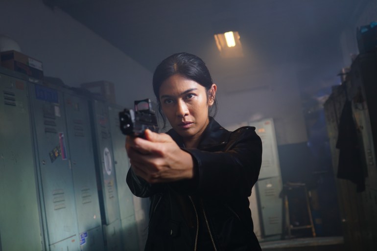 Dian Sastrowardoyo in a still from the film Ratu Adil. She is dressed in black with her hair tied back. She is pointing a gun and looks serious.