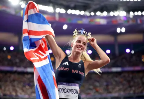 Getty Images Keely Hodgkinson holds a Union Jack flag in one hand and a crown on her head in the other. She is wearing a dark blue vest with Great Britain and her name on. The crowd behind her is blurred.