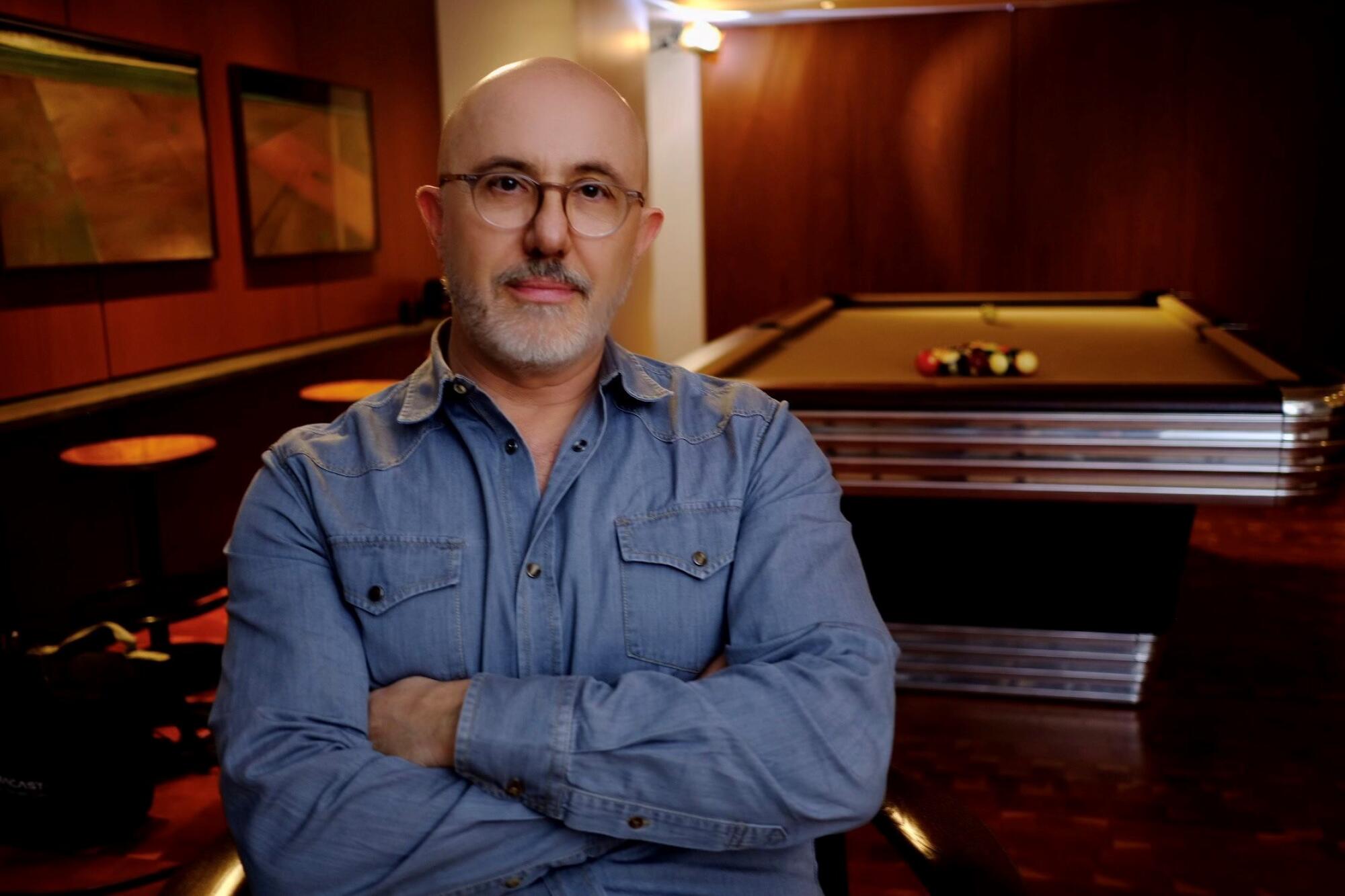 A man folds his arms across his blue shirt and looks straight ahead while sitting in front of a pool table.