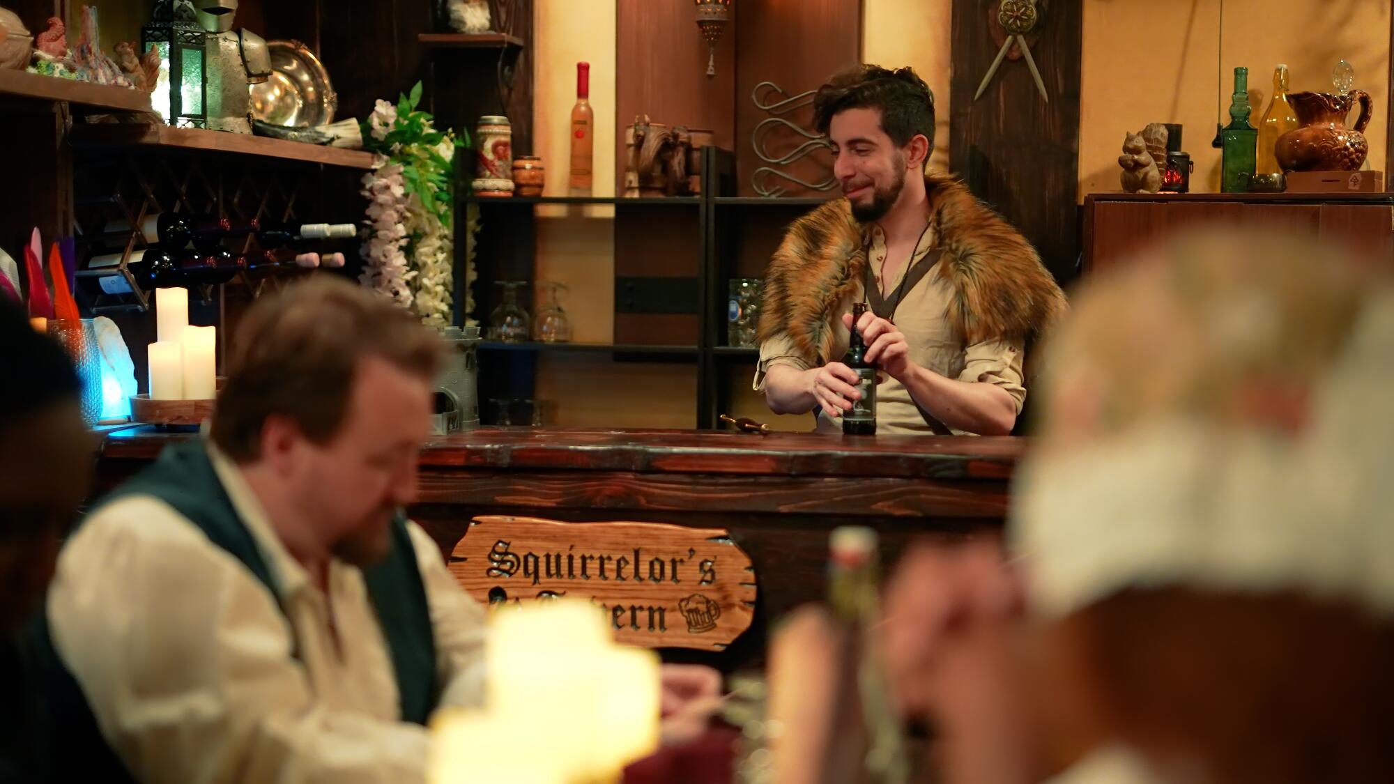 A bearded man in a fantasy costume stands behind the wooden bar in a pub. 