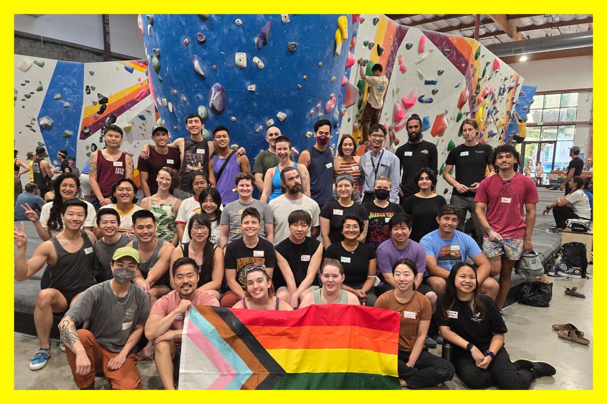 A group of folks pose for a photo with an indoor climbing wall behind them and a pride flag in front of them.