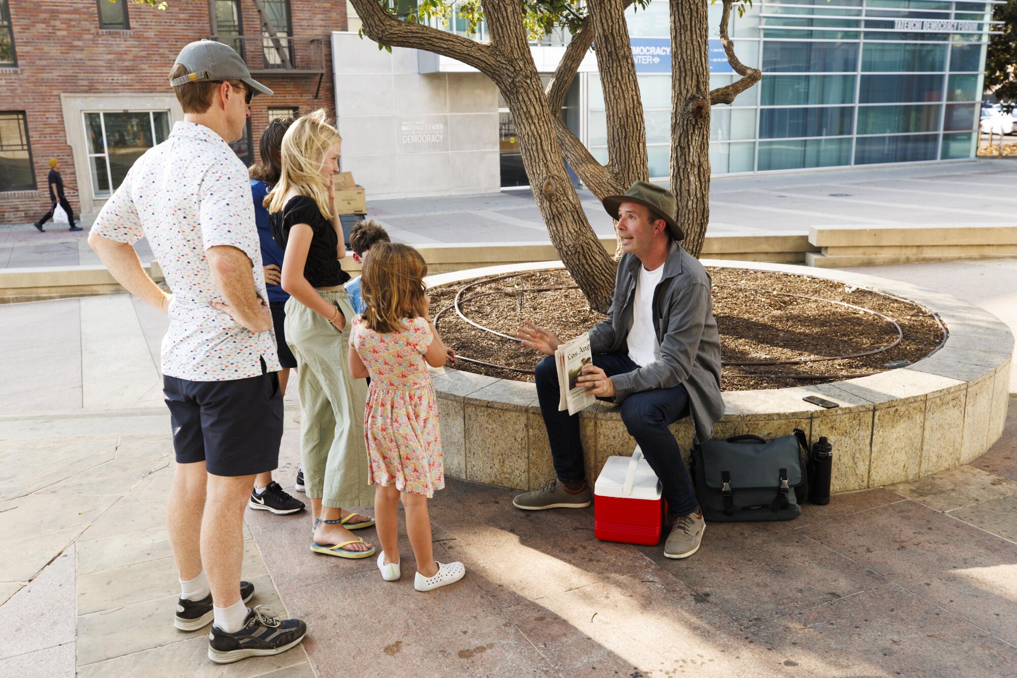 A man sitting on a low wall around a tree talks to a family standing in front of him