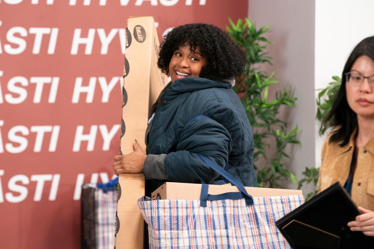 A woman in a blue puffer coat carrying a tall box and two full bags out of a store.