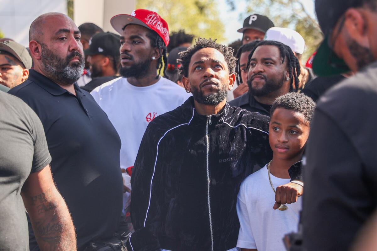 Kendrick Lamar in a dark jacket looking up. He is surrounded in Compton by a crowd of people in casual clothing.