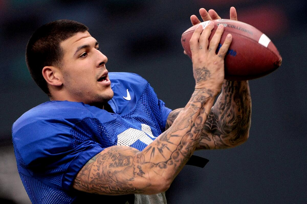 A football player with heavily tattooed arms in a blue jersey and pads, holding a football in his hands near his face.