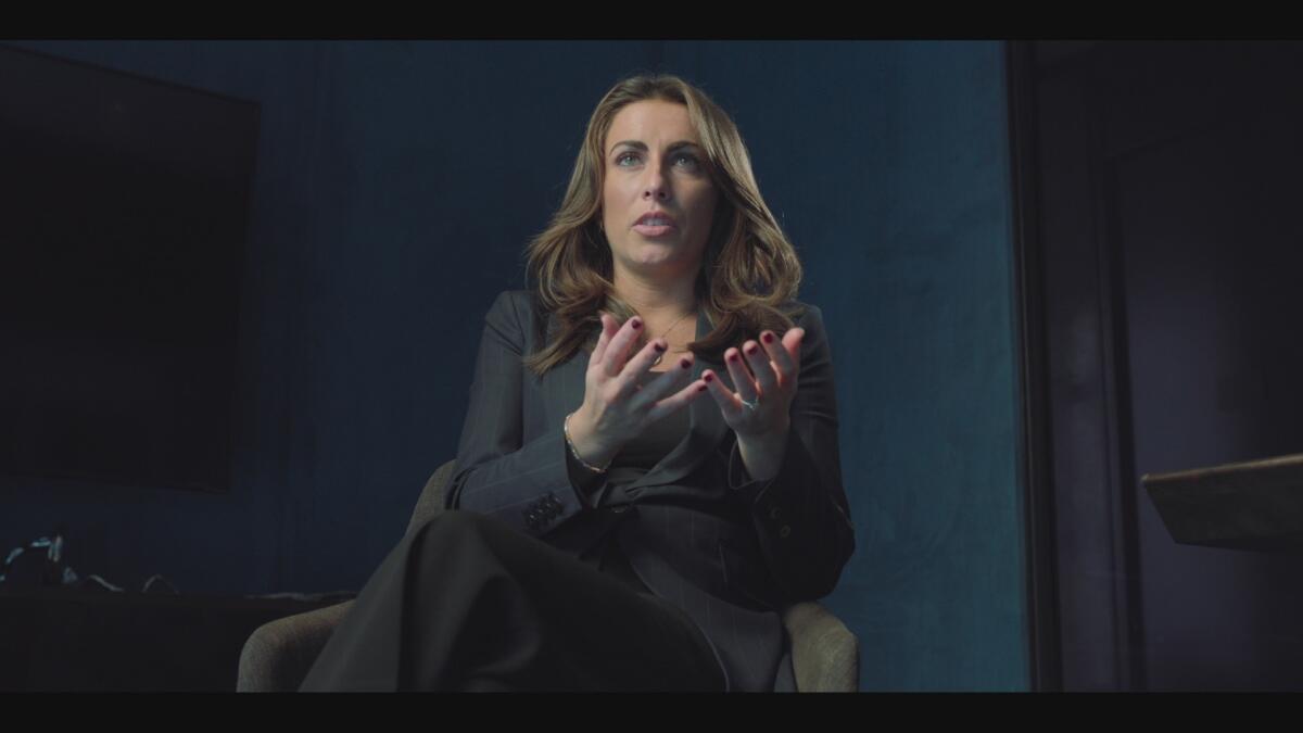 A woman with long hair in a gray suit sits and holds her hands in front of her.