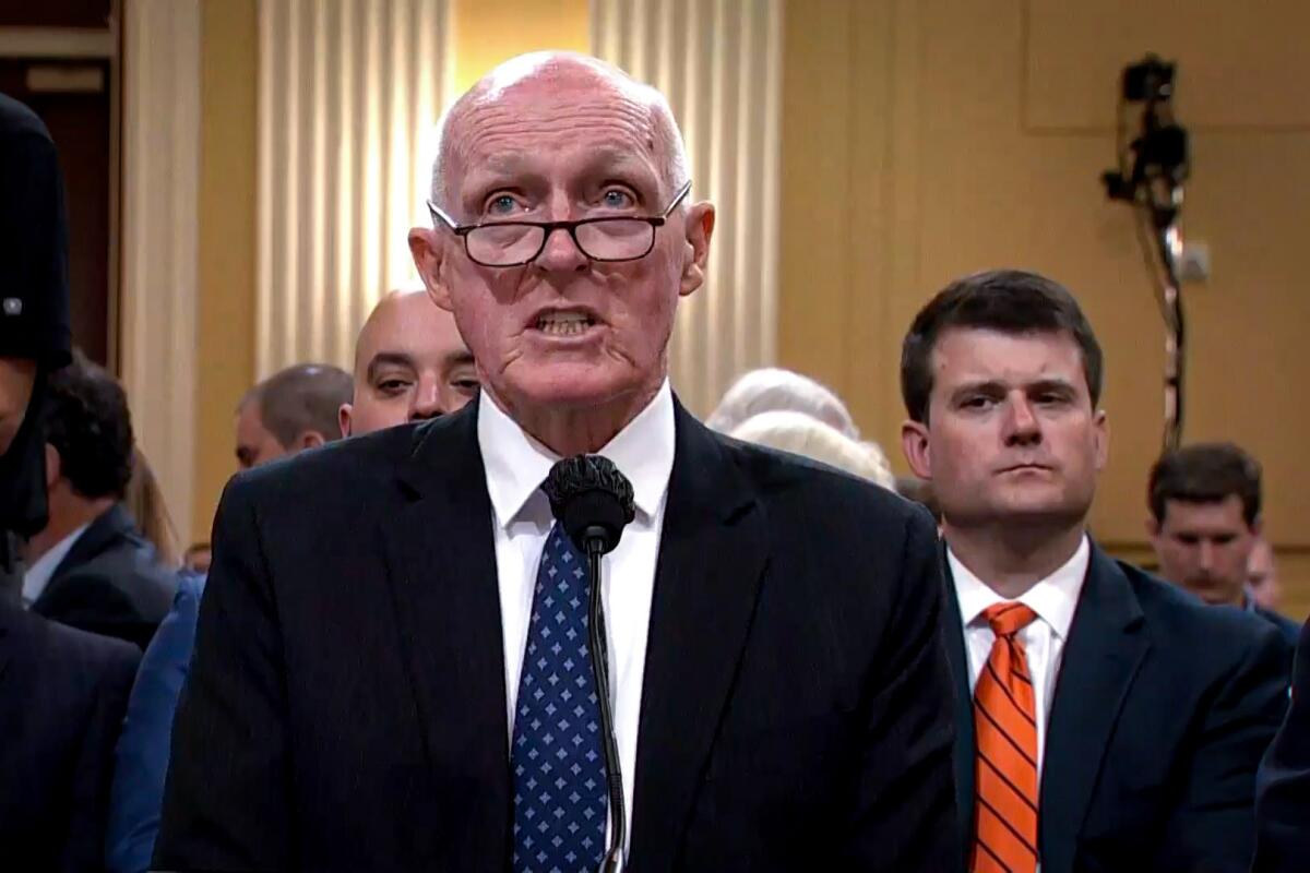 A bald man wearing glasses in a dark suit and blue tie.