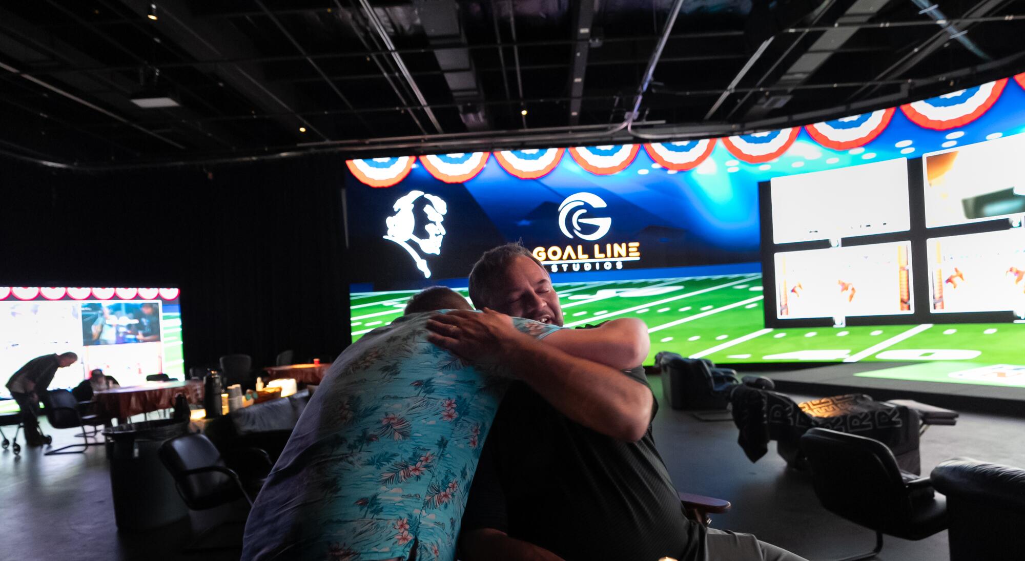 Joe and Jack Madden hug in front of a large screen.