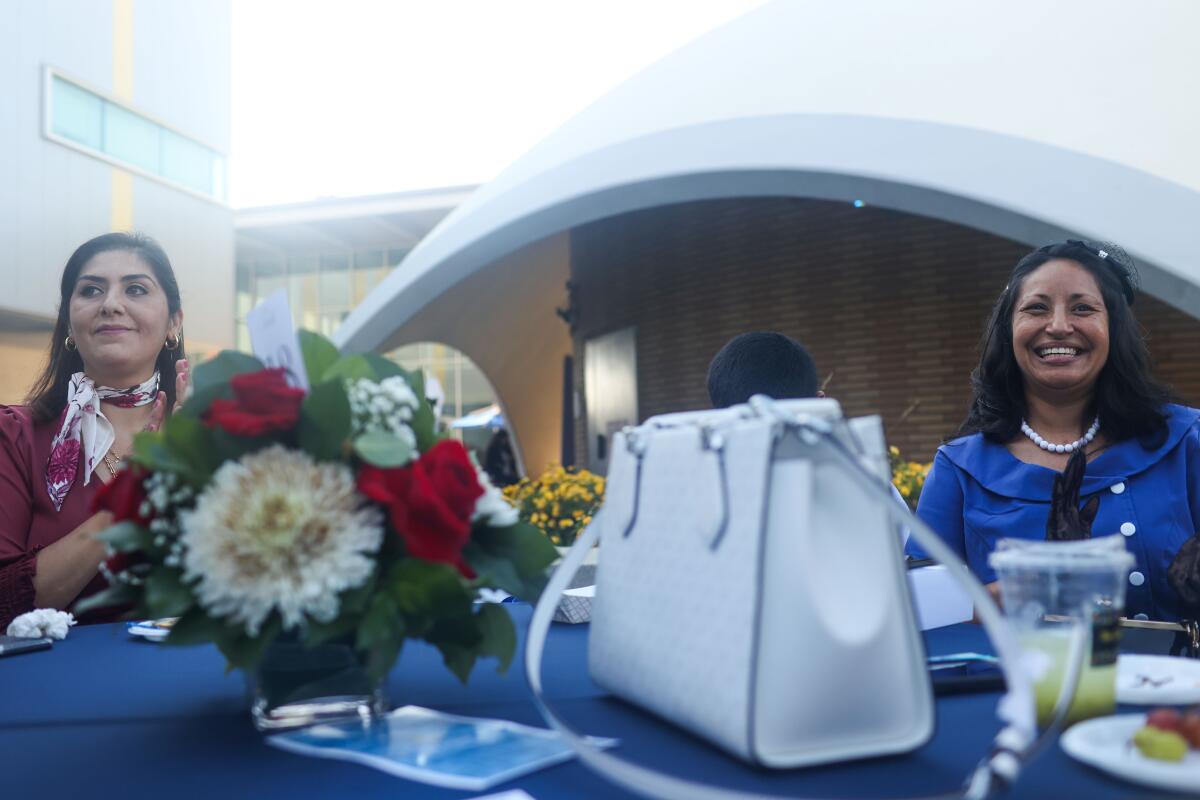 Two women sit behind a table with flowers, a purse and other items on it.