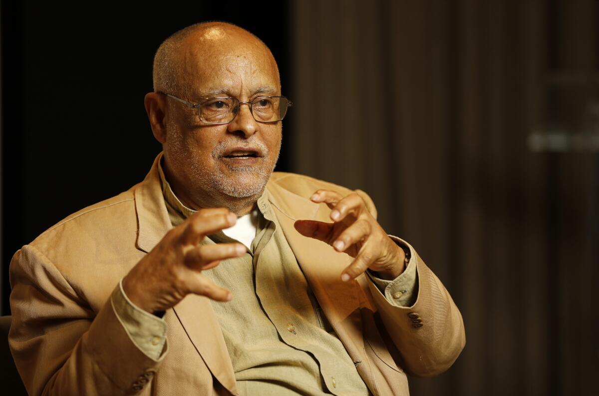 A man in a tan suit speaking and gesturing with his hands