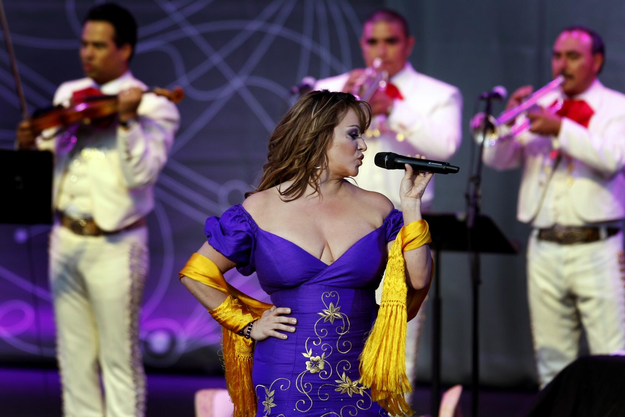 Jenni Rivera, in a low-cut blue dress and yellow shawl, sings onstage backed by mariachi musicians