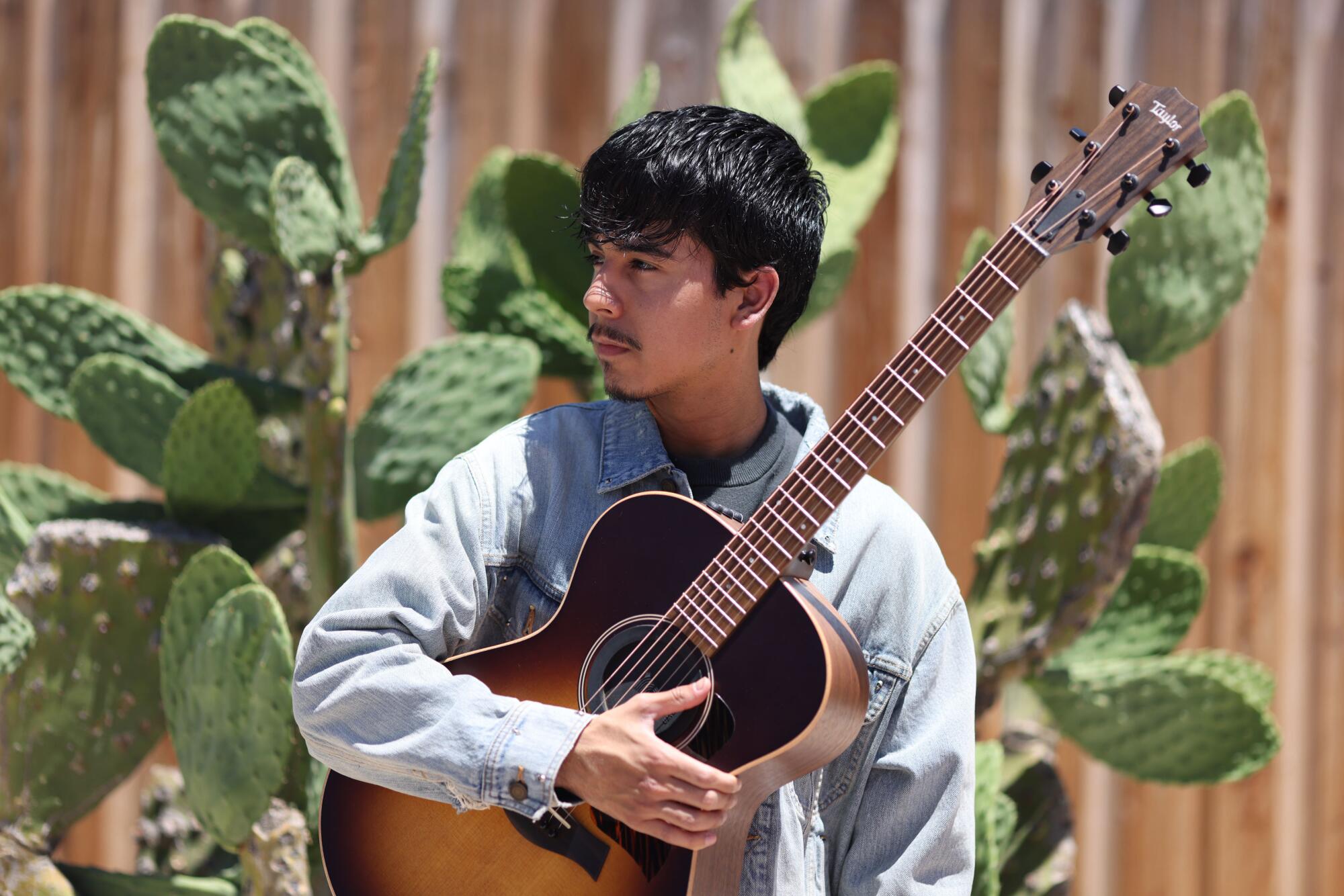 Singer Ivan Cornejo stands outside in front of a large cactus, holding a guitar