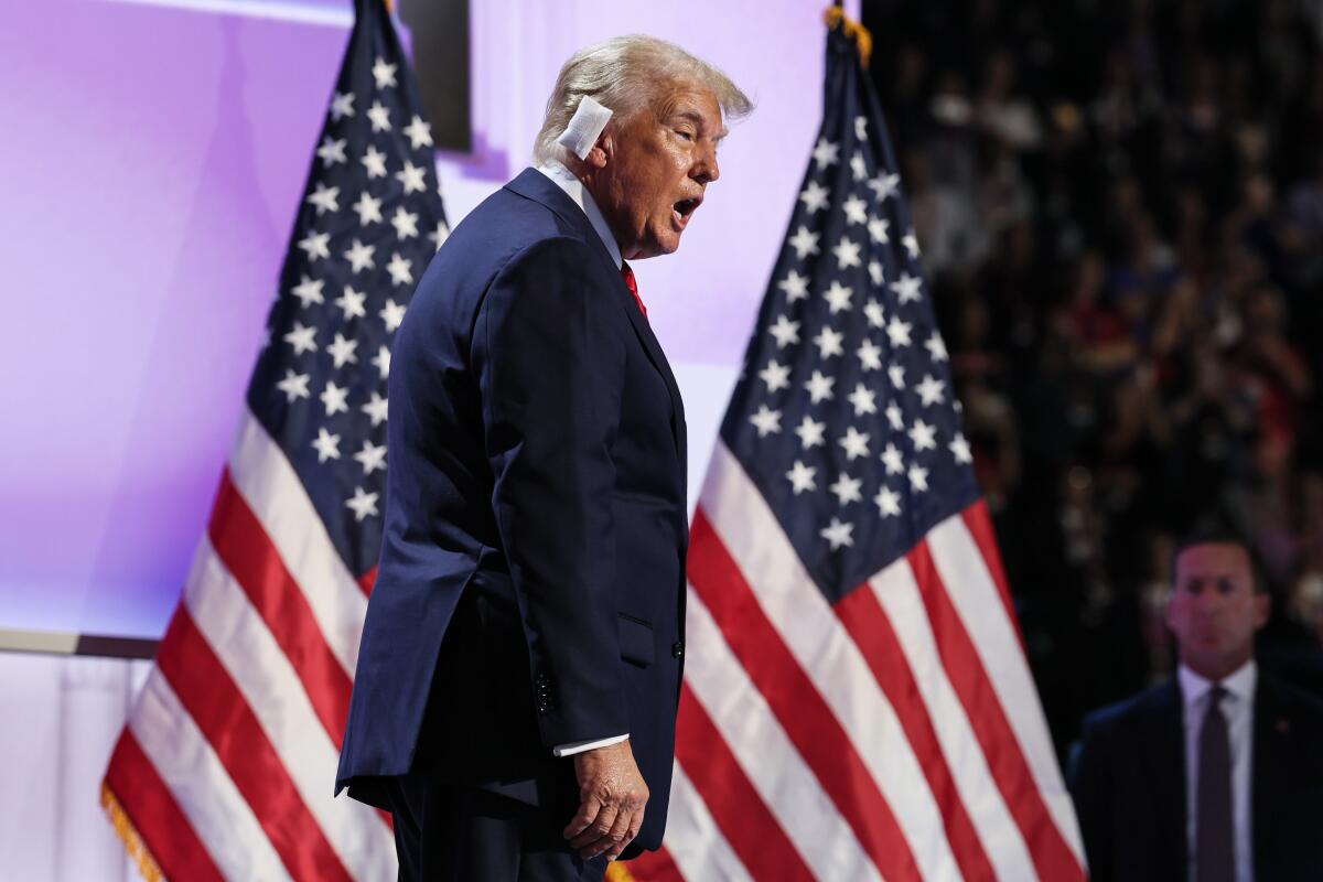 Donald Trump wearing a large white bandage on his ear on a stage with American flags behind him.