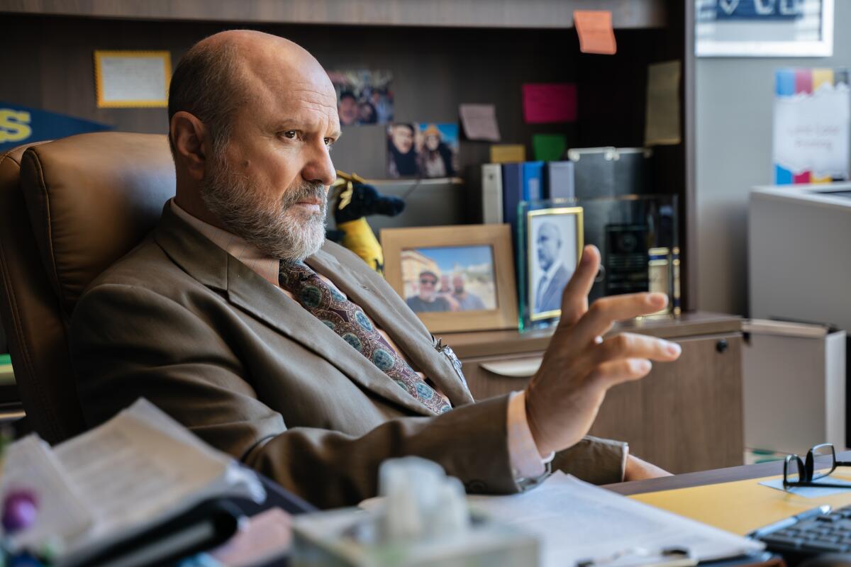 A man in a suit and tie sitting at a desk and leaning back with his hand extended