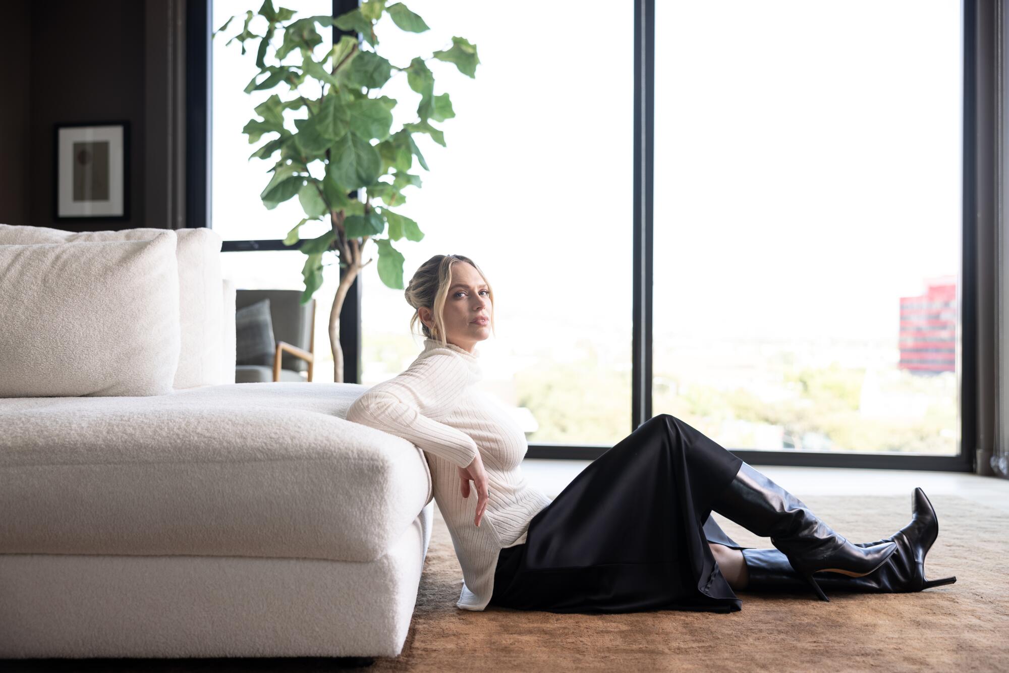 A woman in a white sweater, black skirt and boots leans against a white couch.