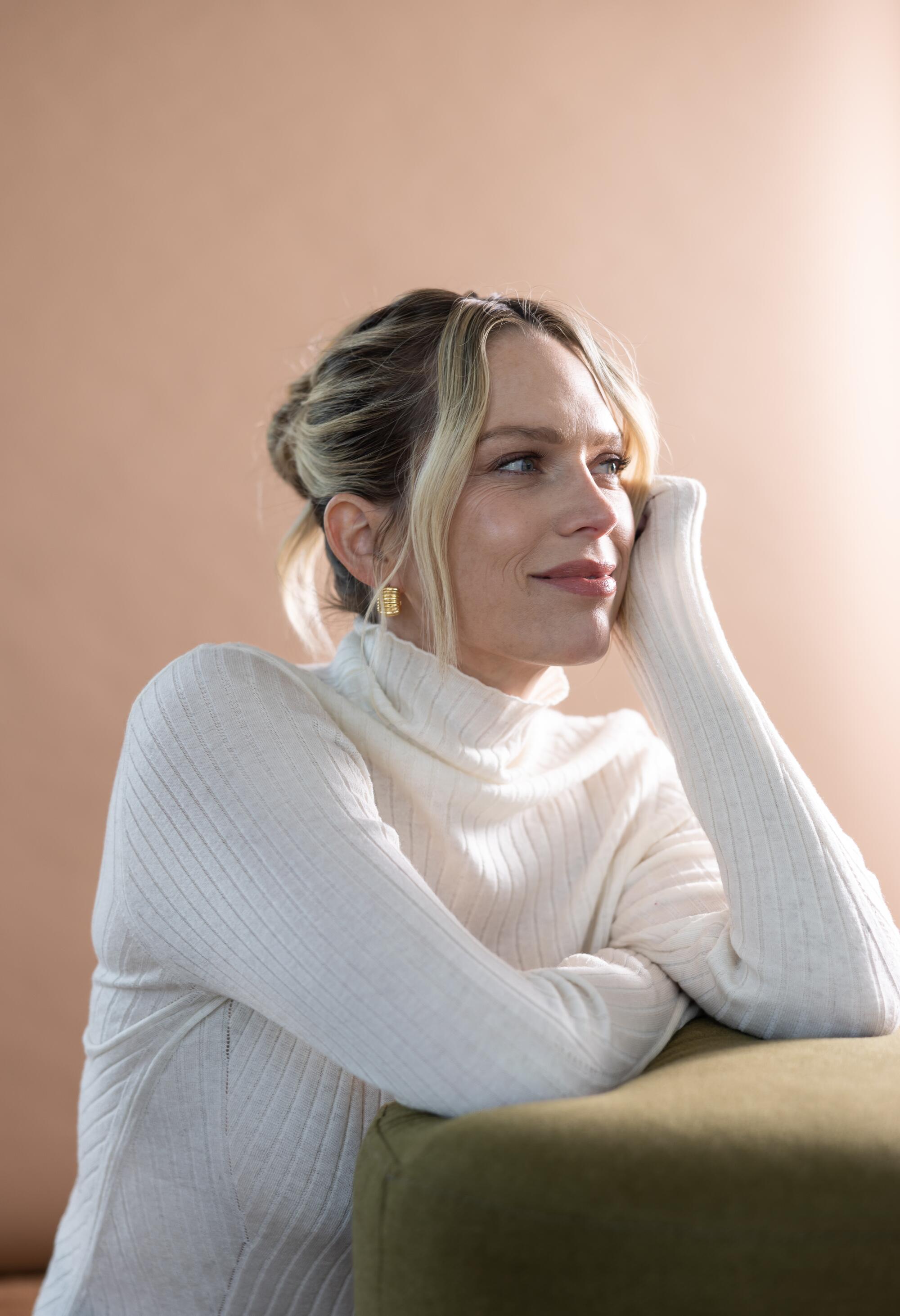 A blonde woman in a white sweater leans against a tan cushion.