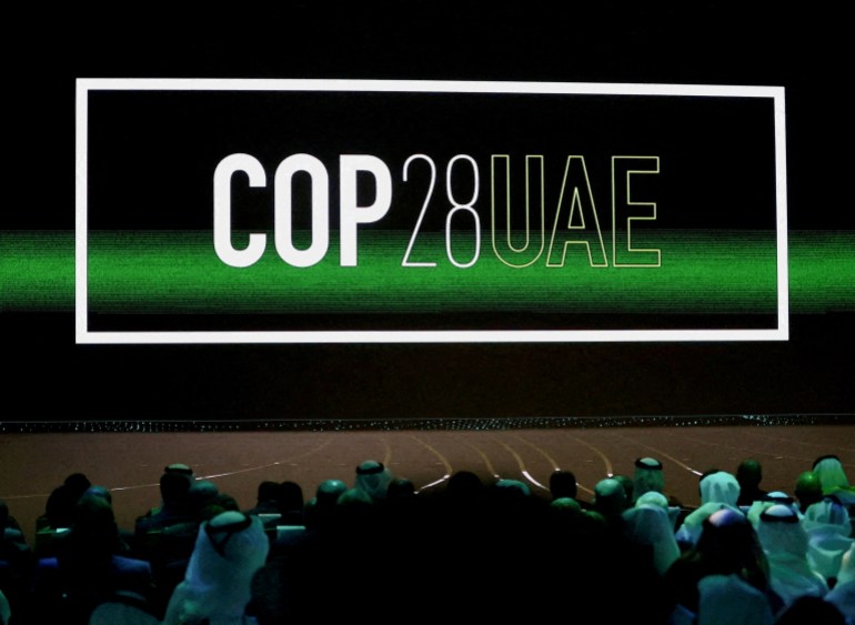 'Cop28 UAE' logo in white is displayed on the screen with a green beam across it, dimly lit audience in the crowd in Abu Dhabi