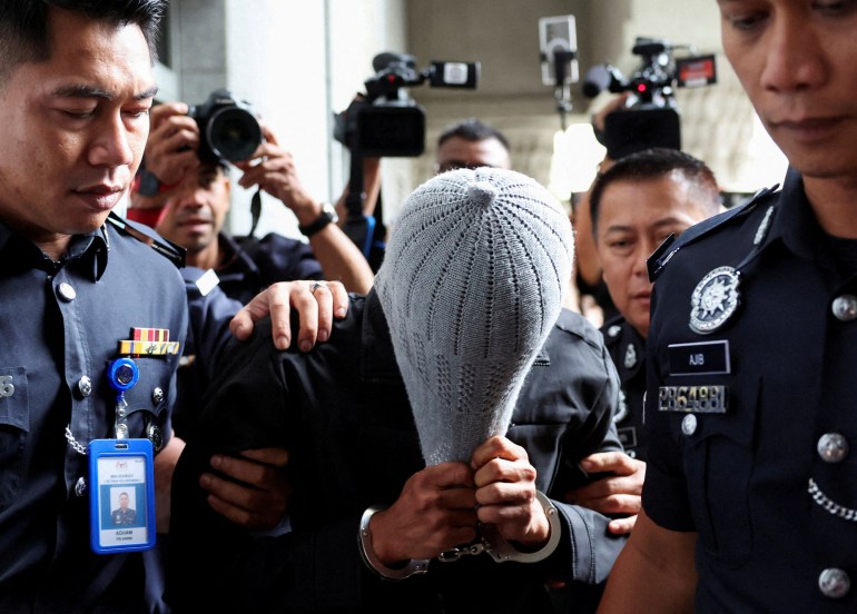 A man linked to Global Ikhwan Service and Business Holdings (GISB) covers his face as he is escorted by police officers after being charged in a court in Putrajaya, Malaysia