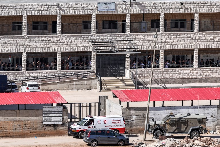 army vehicles and ambulance outside a school where students stand behind bars watching