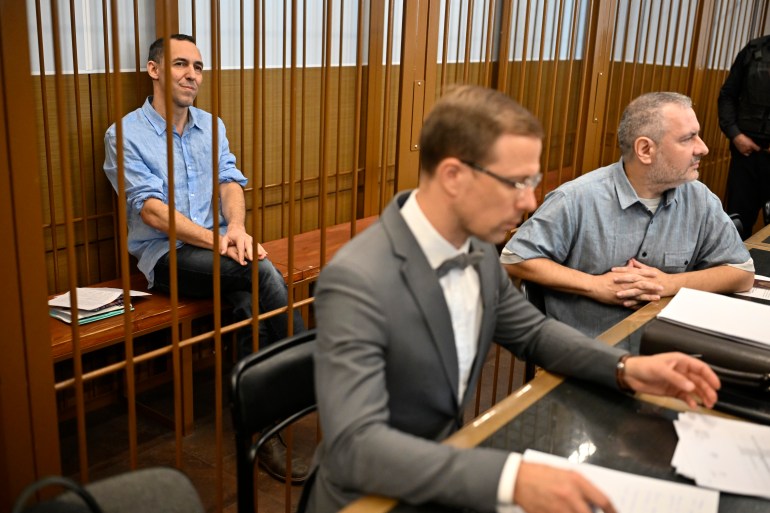 Laurent Vinatier sitting in a caged dock at his trial. He is wearing a pale blue shirt and dark trousers and his hands on his knees. His lawyers are sitting at a desk in front of the cage