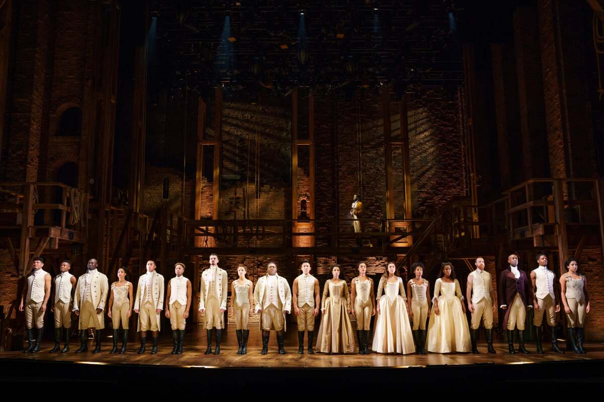 A line of actors in white and beige Colonial costumes prepares to take a bow at the edge of the stage for "Hamilton."