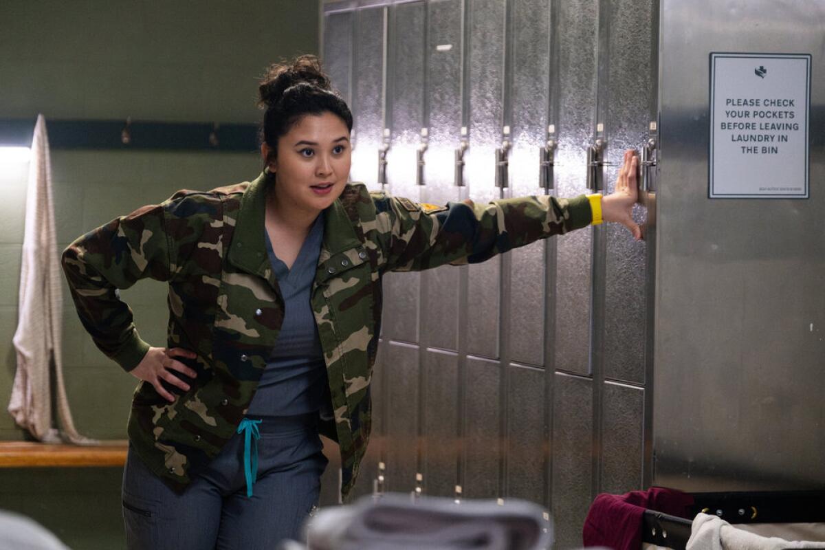 A woman in blue scrubs and a camo jacket leans her hand against a row of gray lockers.