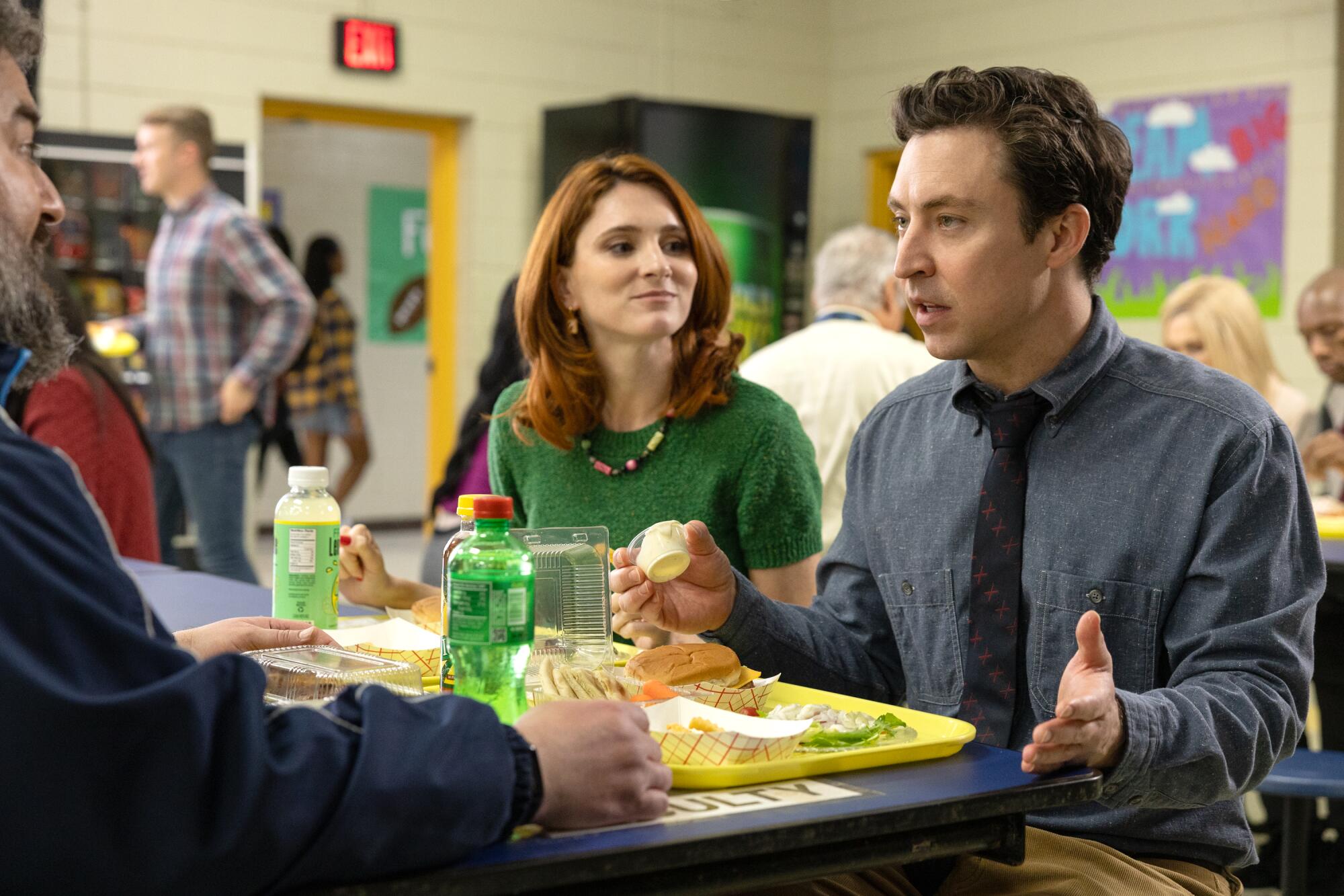 A redheaded woman in a green shirt seated next to a man in a blue shirt and dark tie at a lunch table.