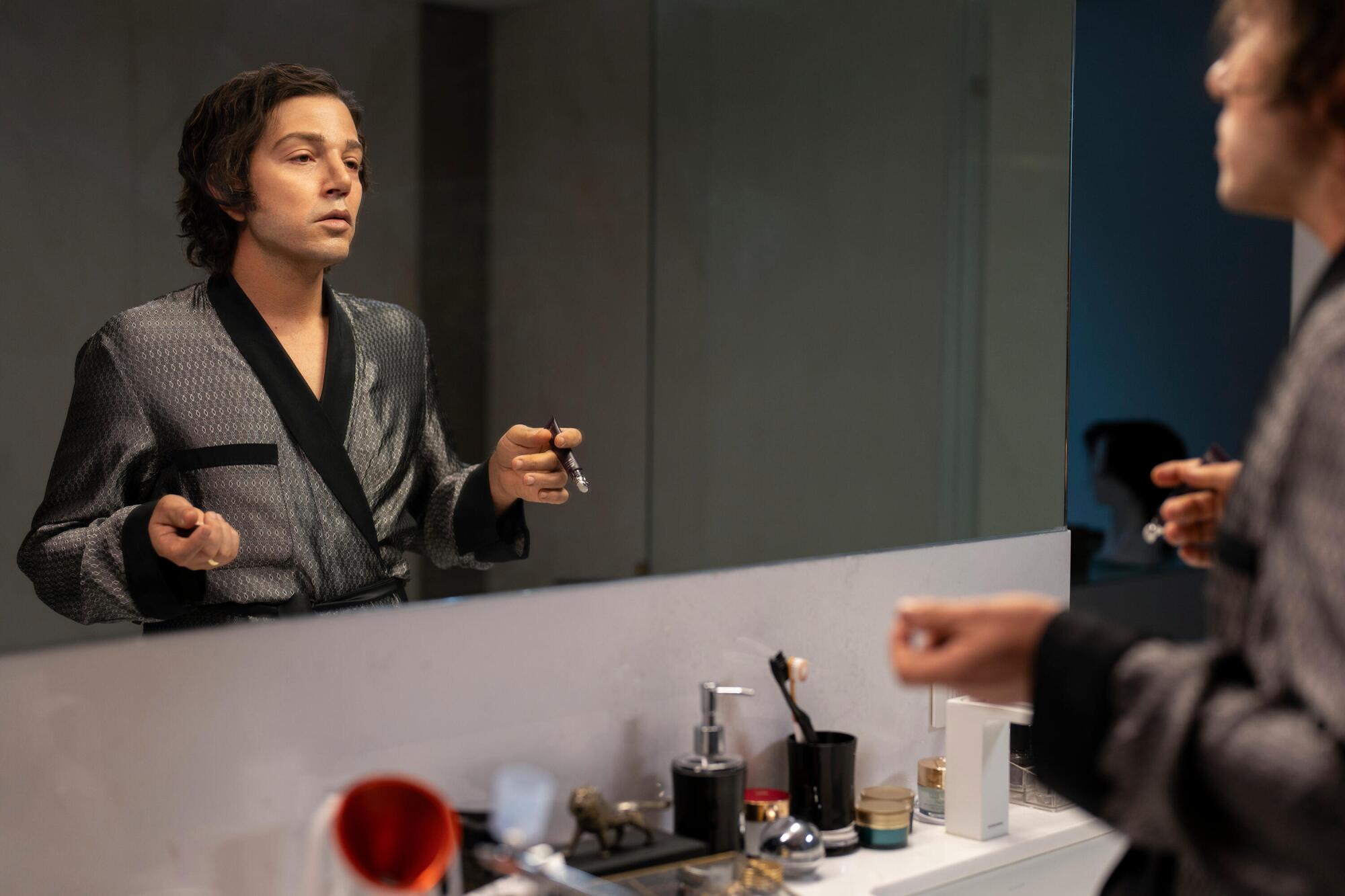 A man in a black and gray robe stares at himself in a mirror.