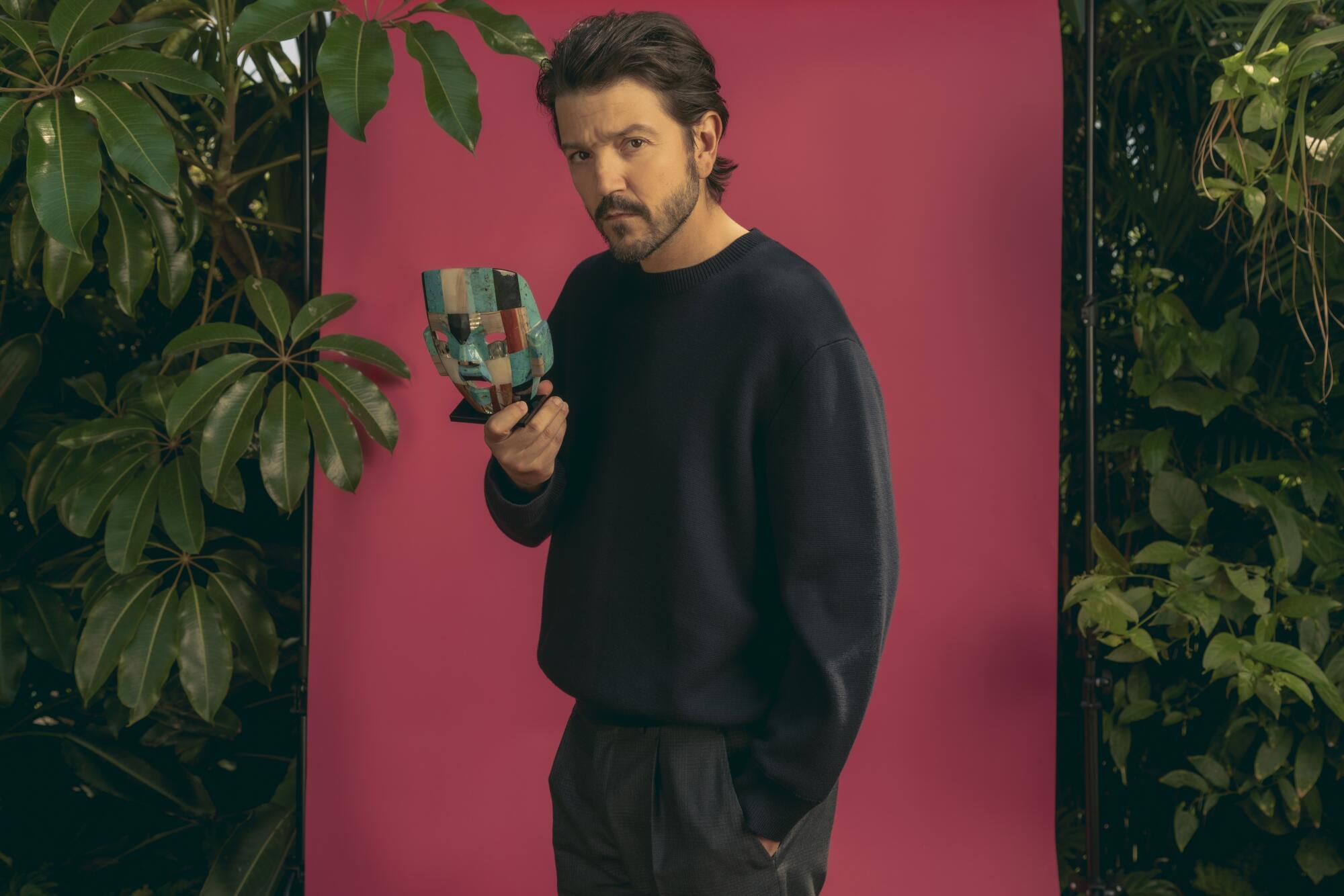 A man in dark clothing stands in front of a pink backdrop and greenery while holding a mask near his face.