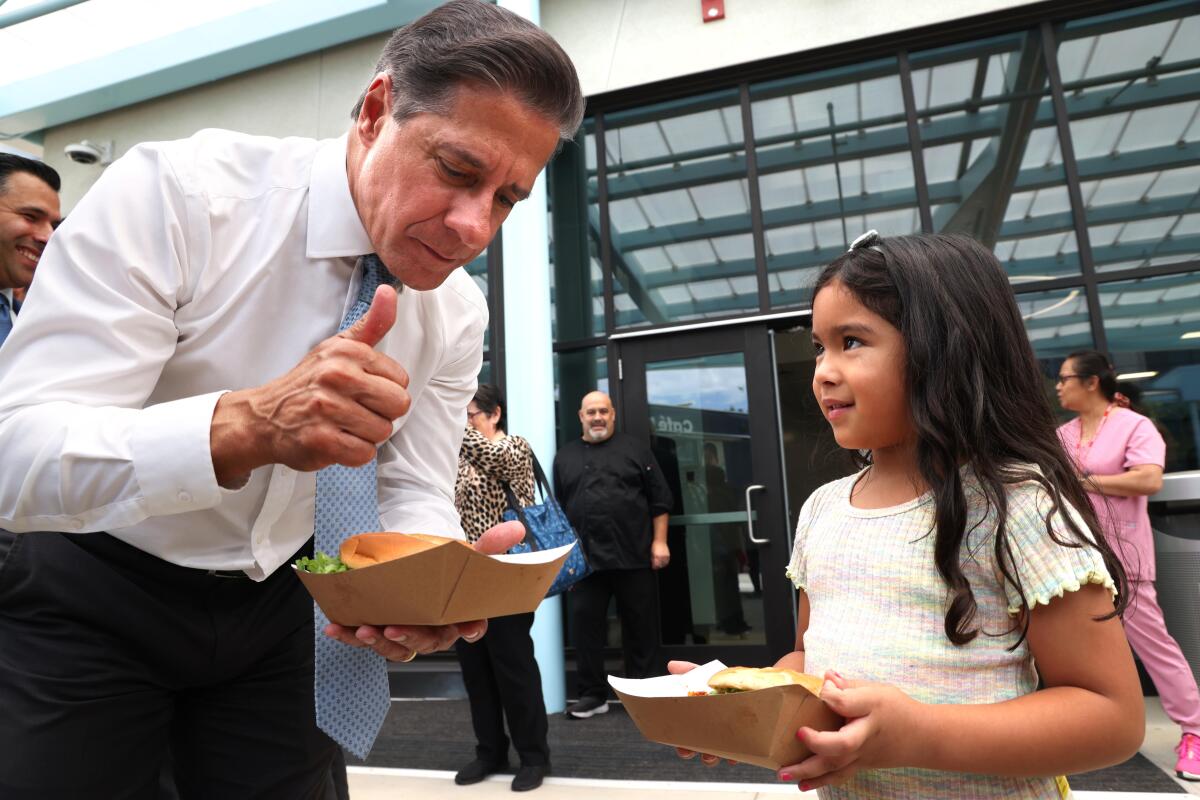 Alberto Carvalho gives a thumbs-up as he and a young girl each hold a fresh meal in a cardboard tray in a school cafeteria