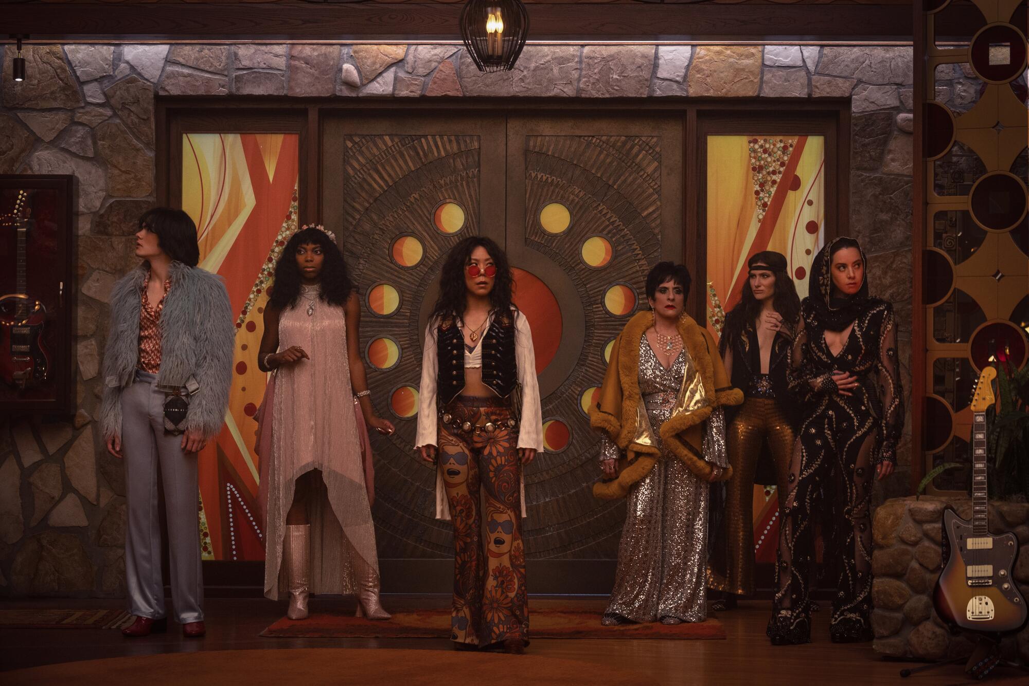 Six people dressed in '70s rock outfits standing in front of a door