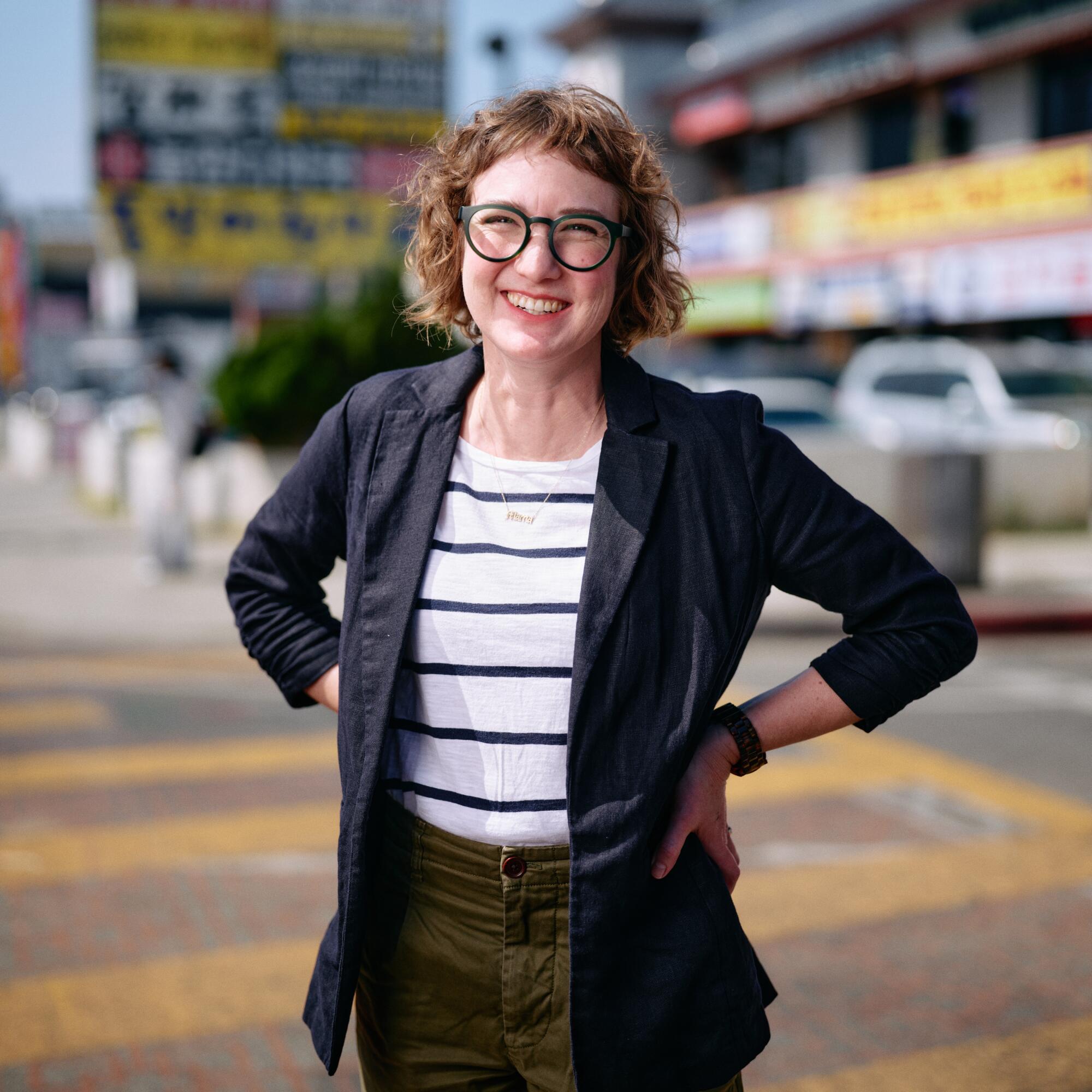 A smiling woman stands with her hands on her hips outdoors.