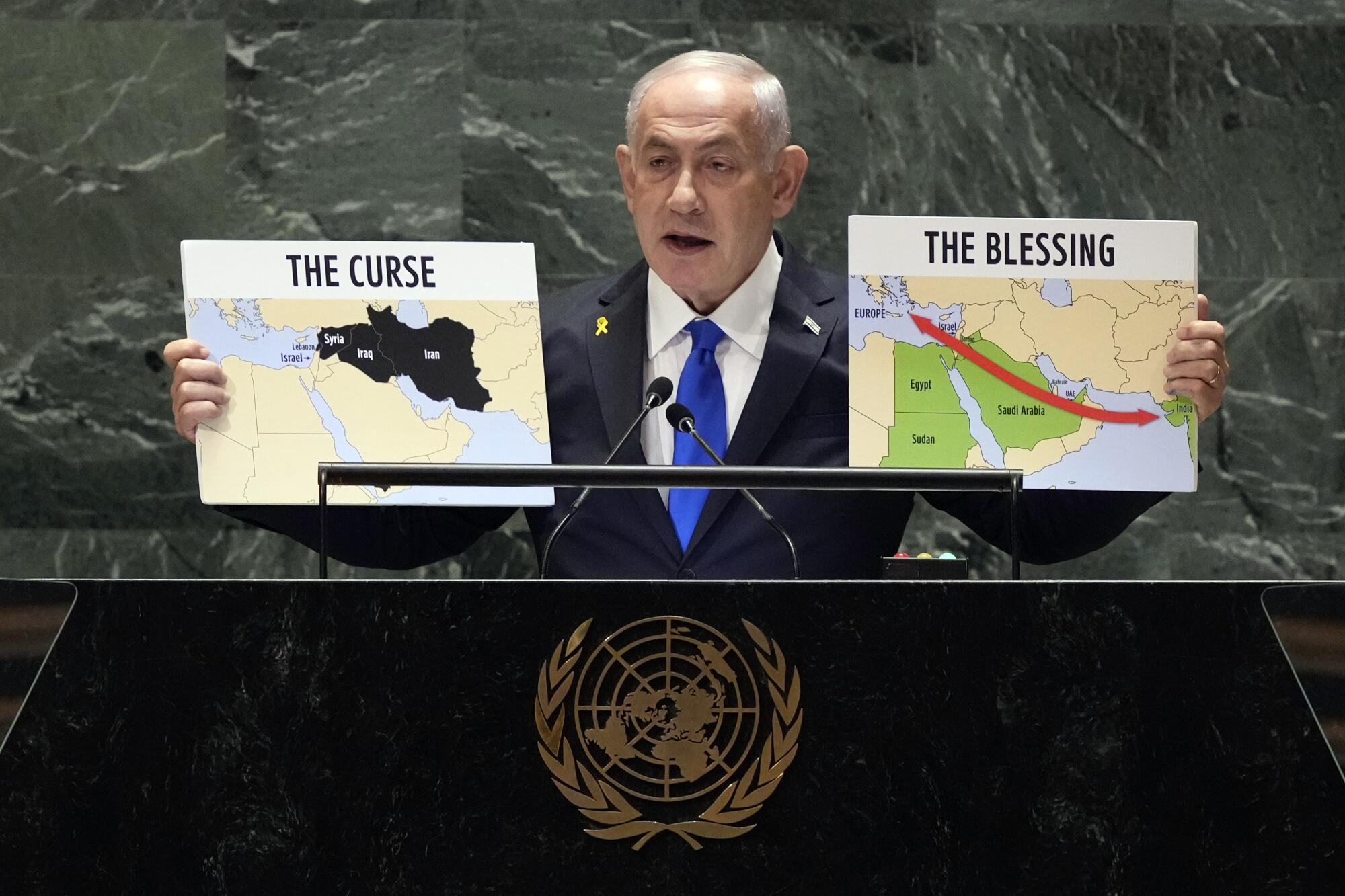 A man in dark suit and blue holds up two posters of maps, one says The Curse and the other titled The Blessing