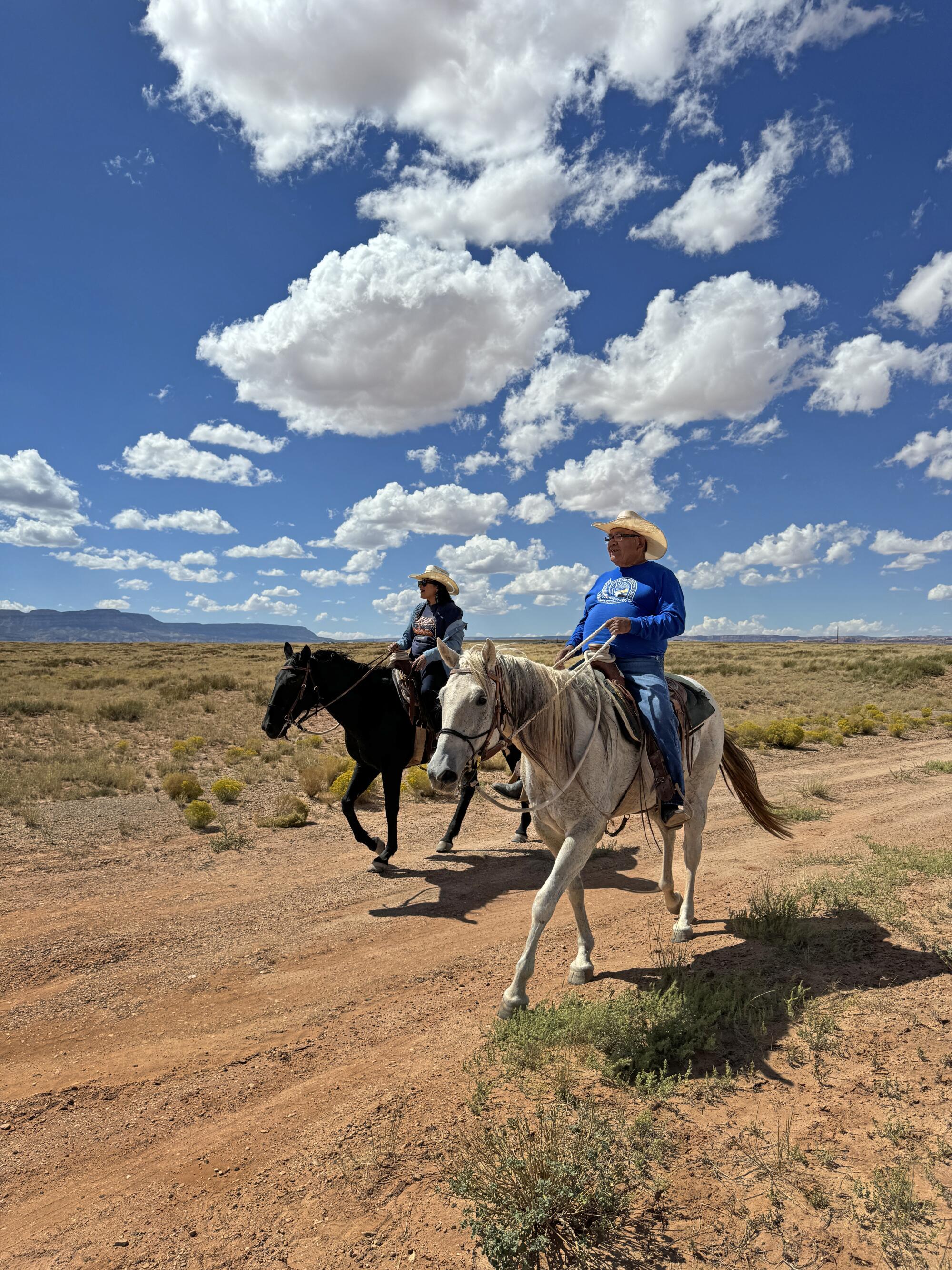 Allie Redhorse Young and her father, Frank Young, ride horses on a dirt trail beneath a big blue sky with puffy white clouds.