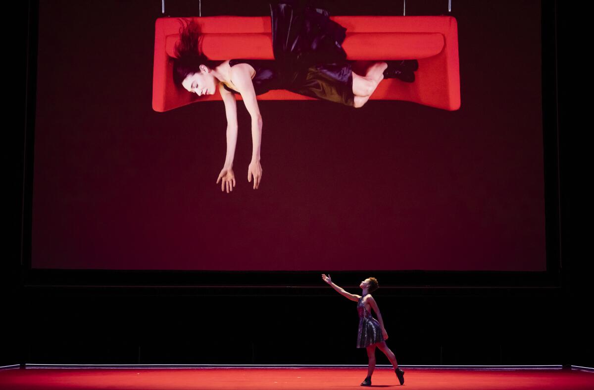 A person onstage reaches up toward a projection of a woman hanging from above, on an upside-down couch, reaching her arms out