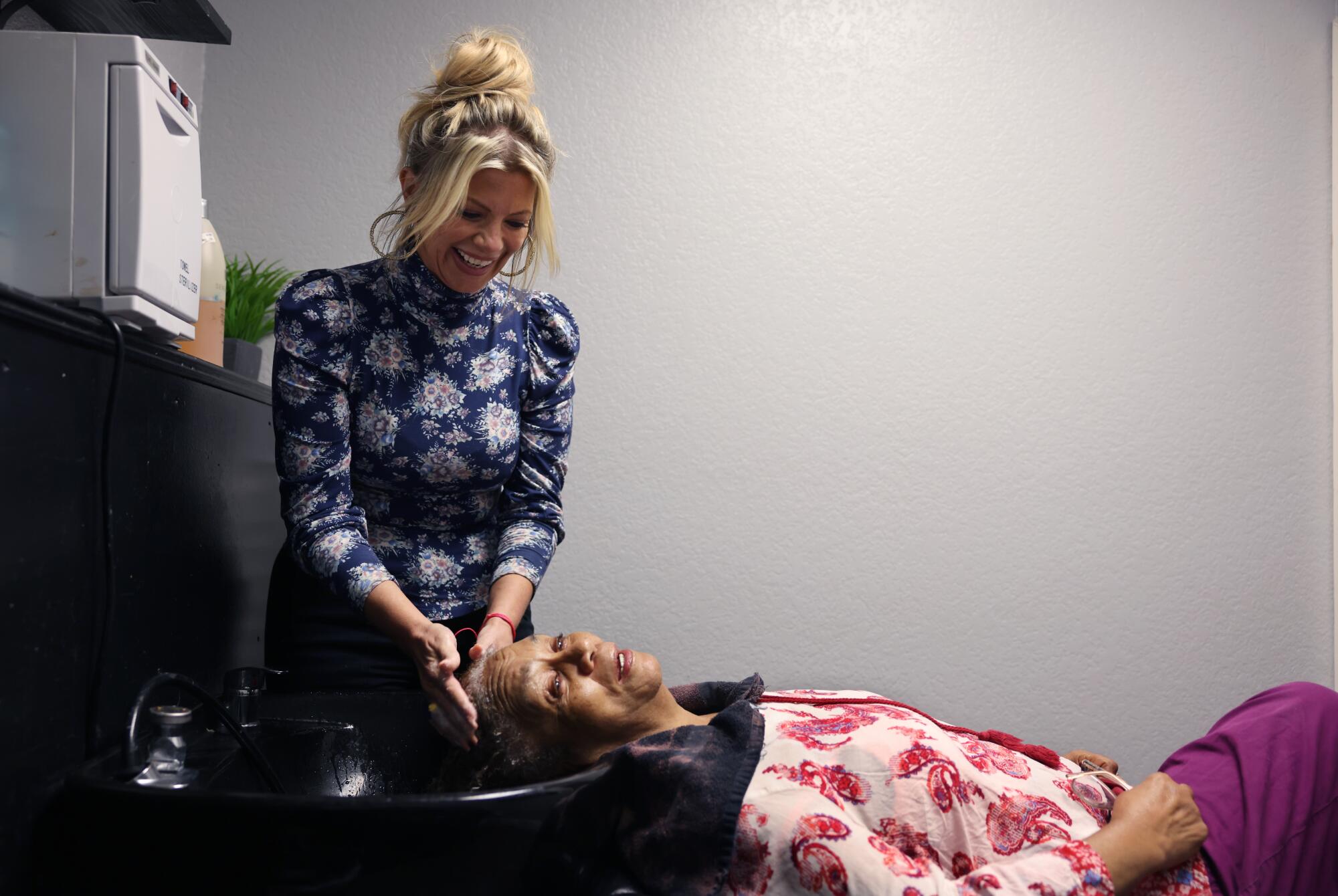 A woman stands over a sink washing the hair of a second woman, who is reclining  