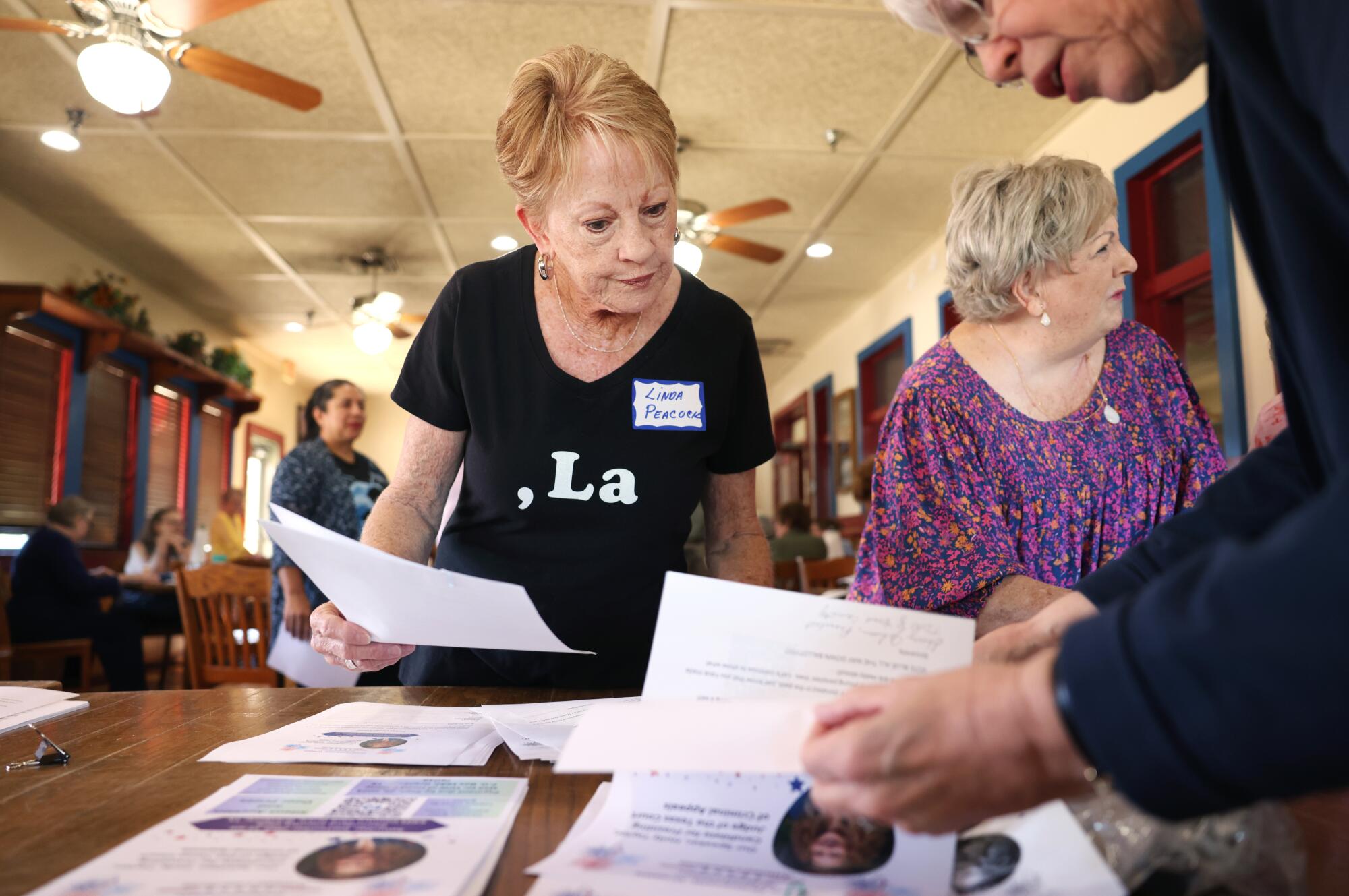 A woman wearing a Kamala T-shirt (written, ", La") joins others looking over pieces of paper on a table   