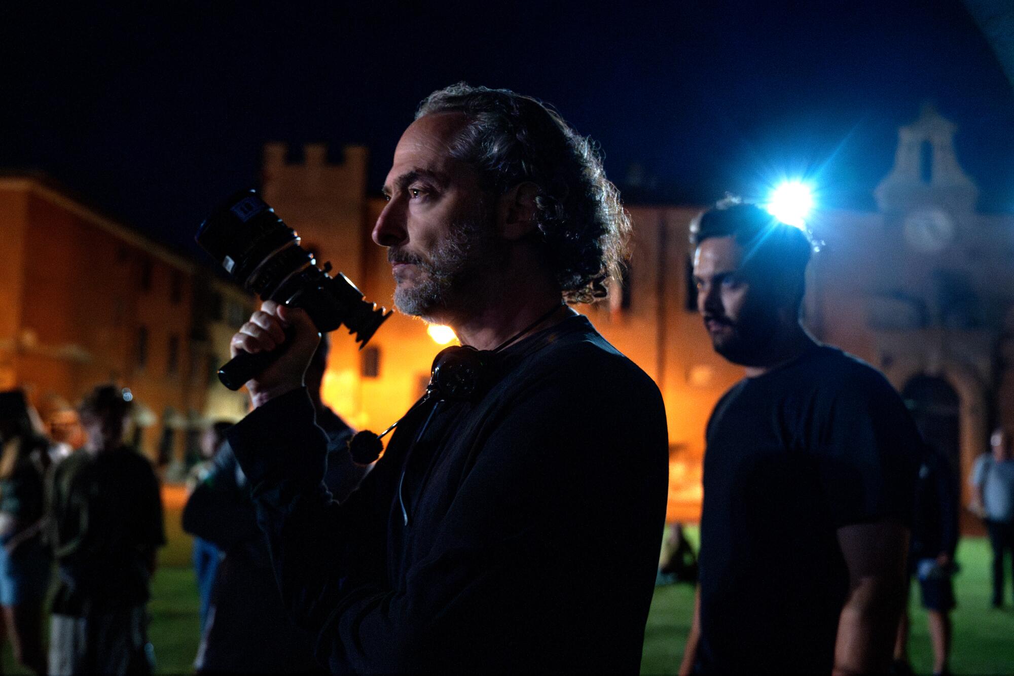 A man holds a camera in one hand while standing outside in the dark.