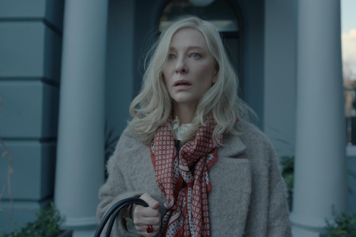 A blond woman in a pale coat and red patterned scarf stands outside a building, looking surprised.